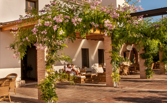 Dos personas sentadas en la terraza de un hotel rural de color blanco rodeado de árboles y flores en un día soleado, un lugar ideal para dormir cerca del Parque Natural Cabárceno.