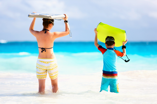 Un niño está en la orilla de la playa con su tabla de surf de color amarilla llevando puesto un bañador azul y a su lado una joven en bikini y short con una tabla de surf de color gris.